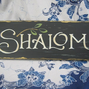 May include: A black wooden sign with gold trim and a white painted inscription that reads "SHALOM". A small green olive branch is painted above the word.