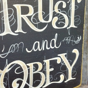 May include: Black wooden sign with white and gold lettering that reads "Trust and Obey".