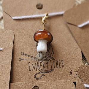 Pode incluir: Uma coleção de pingentes de cogumelos feitos à mão em etiquetas castanhas. Cada pingente apresenta um design de cogumelo com um caule branco e um chapéu castanho ou amarelo, com contas de madeira. As etiquetas estão rotuladas como "EMBERY FIBER".