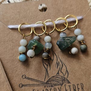 Puede incluir: Marcadores de punto dorados con cuentas de piedras preciosas verdes, blancas y azules. Los marcadores están unidos a una tarjeta marrón con un logotipo. Ideales para proyectos de punto y ganchillo.