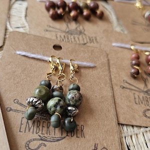 May include: Close-up of handmade stitch markers with gold-colored clasps and various beads in shades of green, brown, and silver. The markers are displayed on brown paper tags with the text "EMBERY FIBER".