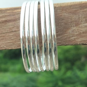 May include: A stack of seven shiny silver bangles. The bracelets are smooth and round, reflecting light. The background is a blurred green, suggesting an outdoor setting. The bangles are resting on a piece of wood.