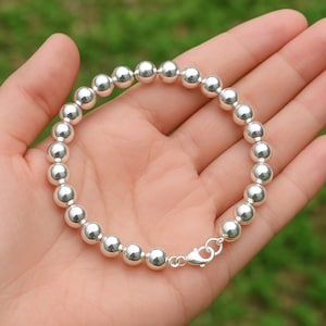 May include: A silver beaded bracelet with a lobster clasp. The bracelet is composed of numerous round, polished silver beads. The bracelet is displayed on a person's open palm against a blurred green background.