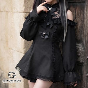 May include: A black long-sleeved dress featuring cold shoulders, a high lace neckline, and a central bow with a heart-shaped charm. The garment includes ruffled accents, lace trim along the hem and sleeves, and a decorative brooch at the collar.