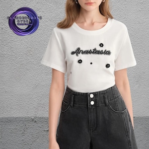 May include: A white t-shirt featuring the name "Anastasia" in black embroidered script, with black flower and dot details. The shirt is worn with black denim shorts. The brand logo is in the top left corner.
