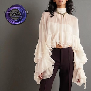 Könnte beinhalten: Eine cremefarbene, transparente Bluse mit Rüschenärmeln und hohem Ausschnitt. Die Bluse wird mit einer dunkelbraunen Hose kombiniert. Das Bild enthält ein Logo mit der Aufschrift "GLAMOURGROVE BYBEA UNISEX FASHION".