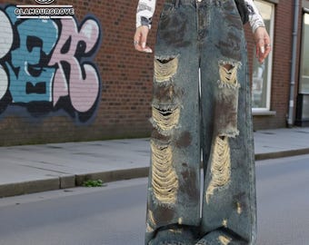 May include: Distressed wide-leg jeans in a faded blue wash with large, ripped details. The jeans have a high waist and a relaxed fit. The image also shows a brick wall with graffiti and the Glamourgrove logo.