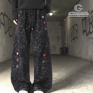 Op de afbeelding: Zwarte broek met wijde pijpen, een gemarmerd patroon en geborduurde rode en blauwe geometrische vormen. De broek heeft een tailleband met trekkoord. Het Glamourgrove-logo is zichtbaar op de achtergrond. Een zwarte trui wordt gedragen.