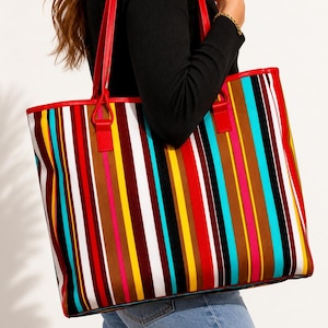 Puede incluir: Un bolso tote rectangular grande con un patrón de rayas verticales vibrantes en tonos rojo, azul brillante, amarillo, blanco, marrón, negro y rosa intenso. El bolso incluye dos correas rojas para el hombro unidas con conectores rectangulares.
