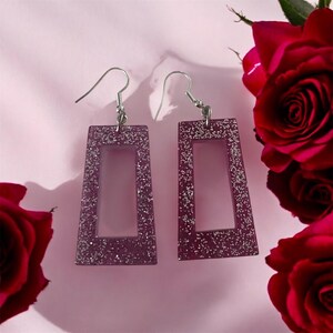 May include: A pair of purple rectangular earrings with silver glitter detailing. The earrings feature a hollow centre and are suspended from silver-coloured fish hook ear wires. The background is a light pink with red roses.