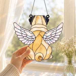 May include: A stained glass suncatcher shaped like a pug dog with angel wings. The pug is tan with black ears, and the wings are made of iridescent white glass. The piece is held by a metal chain and features dark solder lines between glass pieces.