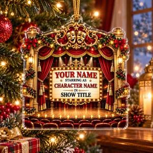 May include: A theater-themed Christmas ornament featuring a stage with red velvet curtains, gold columns, and comedy and tragedy masks. A central marquee sign displays the text: YOUR NAME STARRING AS CHARACTER NAME IN SHOW TITLE.