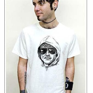 May include: White t-shirt featuring a black and white graphic of a person wearing sunglasses and a hoodie. The person in the image is wearing black pants and has tattoos on their arms. The person is also wearing a black choker and wristband.