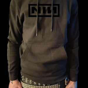 May include: Black hoodie featuring the letters "NIN" within a black rectangle on the chest. The hoodie has a front pocket and drawstrings. The person is wearing a studded belt and rings.