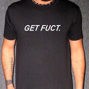 May include: Black t-shirt with the text "GET FUCT." in white, centered on the chest. The shirt has short sleeves and a crew neck. The person is wearing a black necklace with silver spikes.