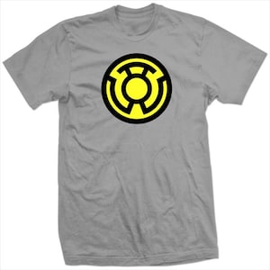 Op de afbeelding: Grijs T-shirt met een cirkelvormig ontwerp met een gele kern en een zwarte omtrek. Het ontwerp is een gestileerd symbool. Het shirt is gemaakt van zacht materiaal en heeft korte mouwen.