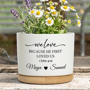 May include: A white ceramic planter with a wooden base, filled with daisies and lavender. The planter has the text "we love because he first loved us 1 John 4:19" and the names "Maya" and "Samuel".
