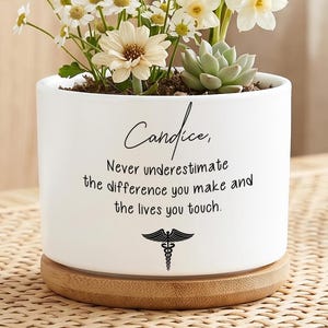 Op de afbeelding: Een witte keramische plantenbak met een houten basis, gevuld met een verscheidenheid aan bloemen en een vetplant. De plantenbak heeft de naam "Candice" en een motiverende quote erop gedrukt, samen met een medisch symbool.