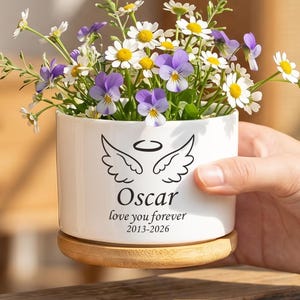 以下が含まれることがあります： 木製の台座が付いた白いセラミックプランター。 デイジーと紫色のパンジーが植えられています。 プランターには天使の羽と「Oscar」の名前と「love you forever 2013-2026」の文字が書かれています。