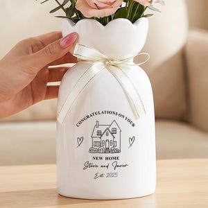 Può includere: Un vaso in ceramica bianca con bordo smerlato e un nastro color crema legato attorno al collo. Il vaso presenta un disegno a linee nere di una casa con il testo "CONGRATULATIONS ON YOUR NEW HOME Stevie and Javier Est. 2025" e due cuori.