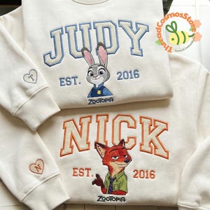 two white sweatshirts with the characters Judy and Nick from the animated film "Zootopia" embroidered on them. The sweatshirts are displayed on a white surface, with the top one having the name "JUDY" and the bottom one having the name "NICK".