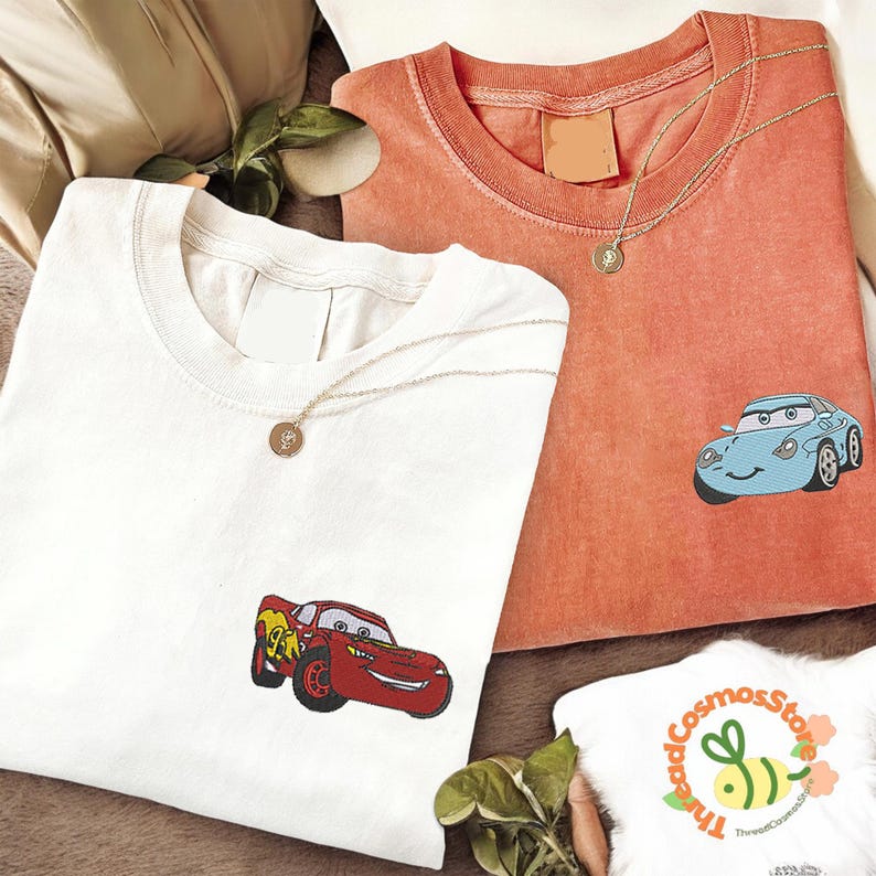 a collection of clothing items, including two t-shirts with cartoon car designs, a necklace, and a small white item with a logo.