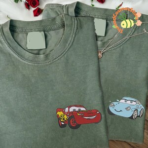 two green t-shirts with cartoon car designs, including a red car and a blue car, embroidered on the front.
