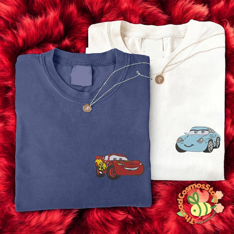 two t-shirts with cartoon car designs, one blue and one white, laid out on a red furry surface.