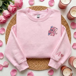 a pink sweatshirt with a cartoon character on the front, placed on a woven placemat. Surrounding the sweatshirt are pink roses, candles, and a wicker basket.