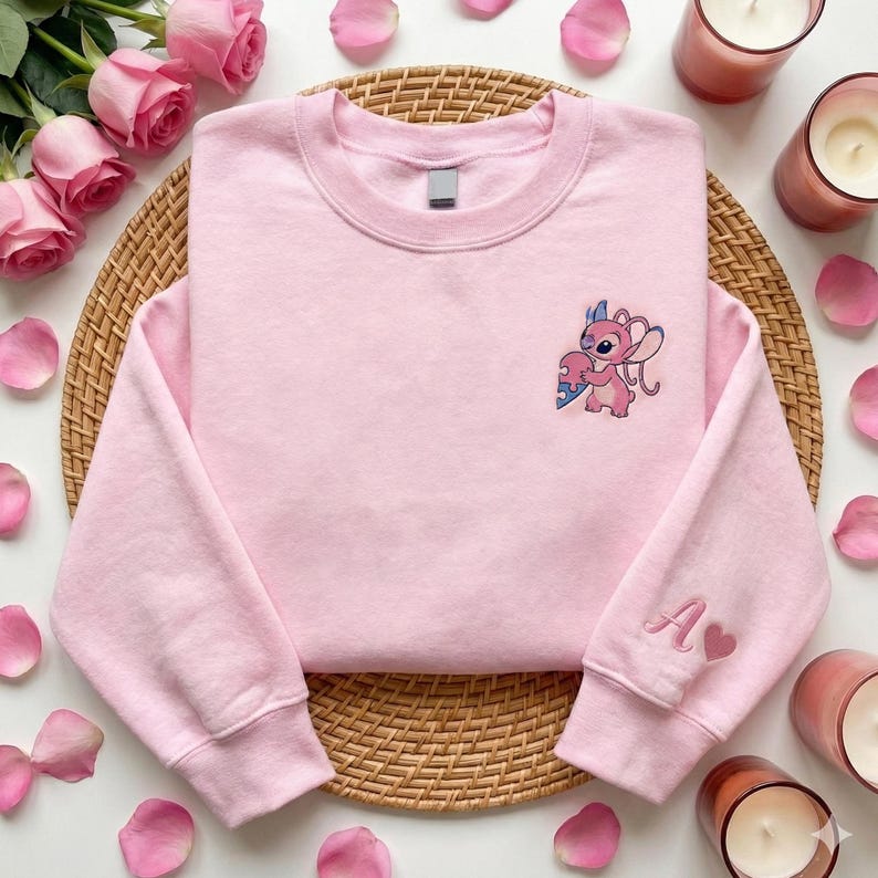 a pink sweatshirt with a cartoon character on the front, placed on a woven placemat. Surrounding the sweatshirt are pink roses, candles, and a wicker basket.