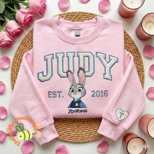 A pink sweatshirt with the word "JUDY" and a cartoon rabbit character is displayed on a woven placemat, surrounded by pink roses and candles.
