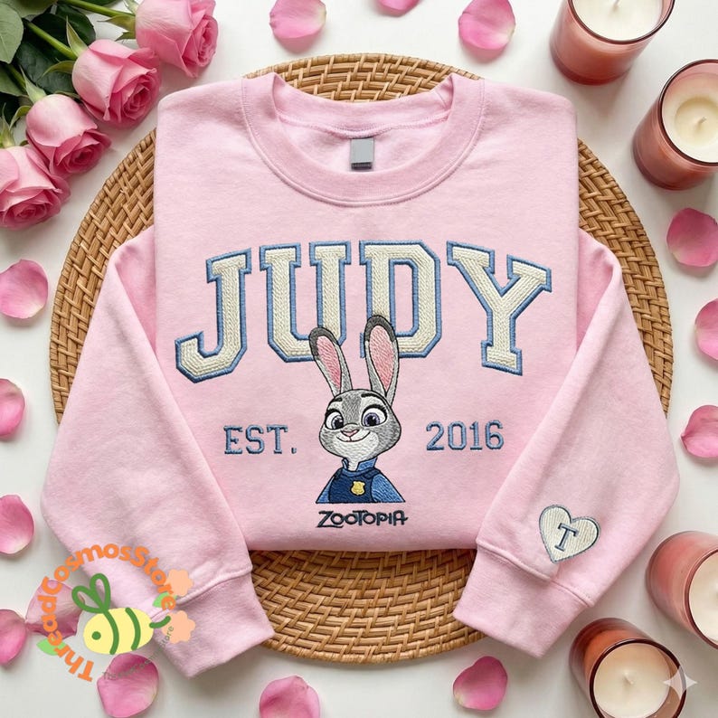 A pink sweatshirt with the word "JUDY" and a cartoon rabbit character is displayed on a woven placemat, surrounded by pink roses and candles.