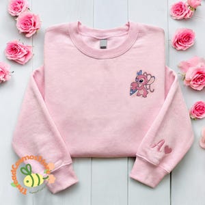 A pink sweatshirt with a cartoon character on the front, placed on a white wooden surface surrounded by pink roses.