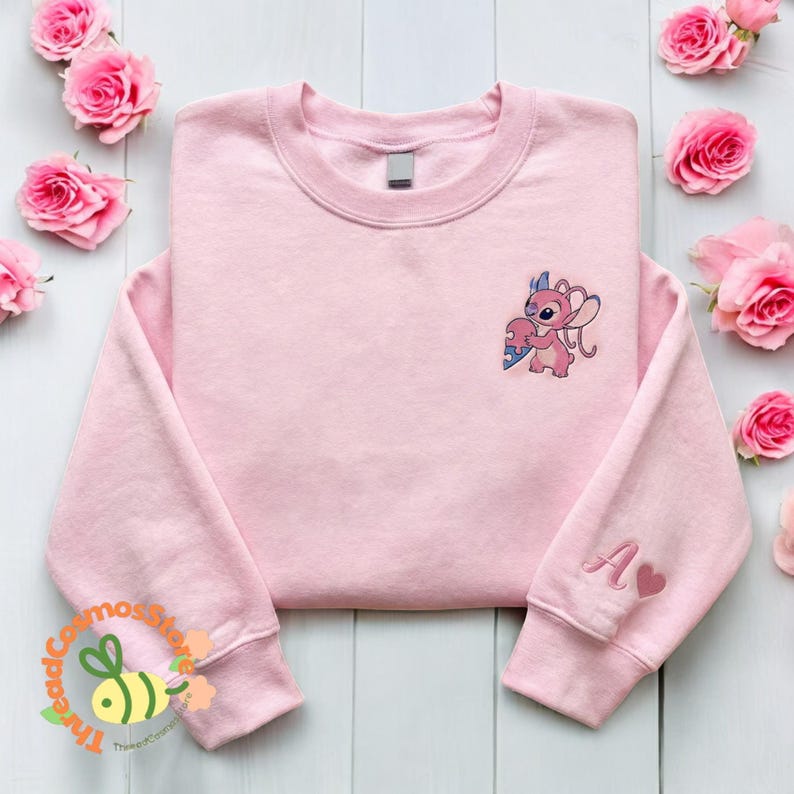 A pink sweatshirt with a cartoon character on the front, placed on a white wooden surface surrounded by pink roses.