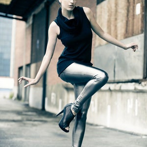 May include: A woman in a black sleeveless top and silver metallic leggings poses outdoors. She wears black high-heeled shoes. Her hair is styled in a bun with a dark flower accessory. The background features brick and concrete.