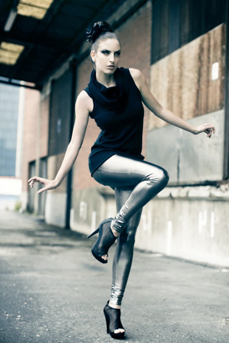 May include: A woman in a black sleeveless top and silver metallic leggings poses outdoors. She wears black high-heeled shoes. Her hair is styled in a bun with a dark flower accessory. The background features brick and concrete.