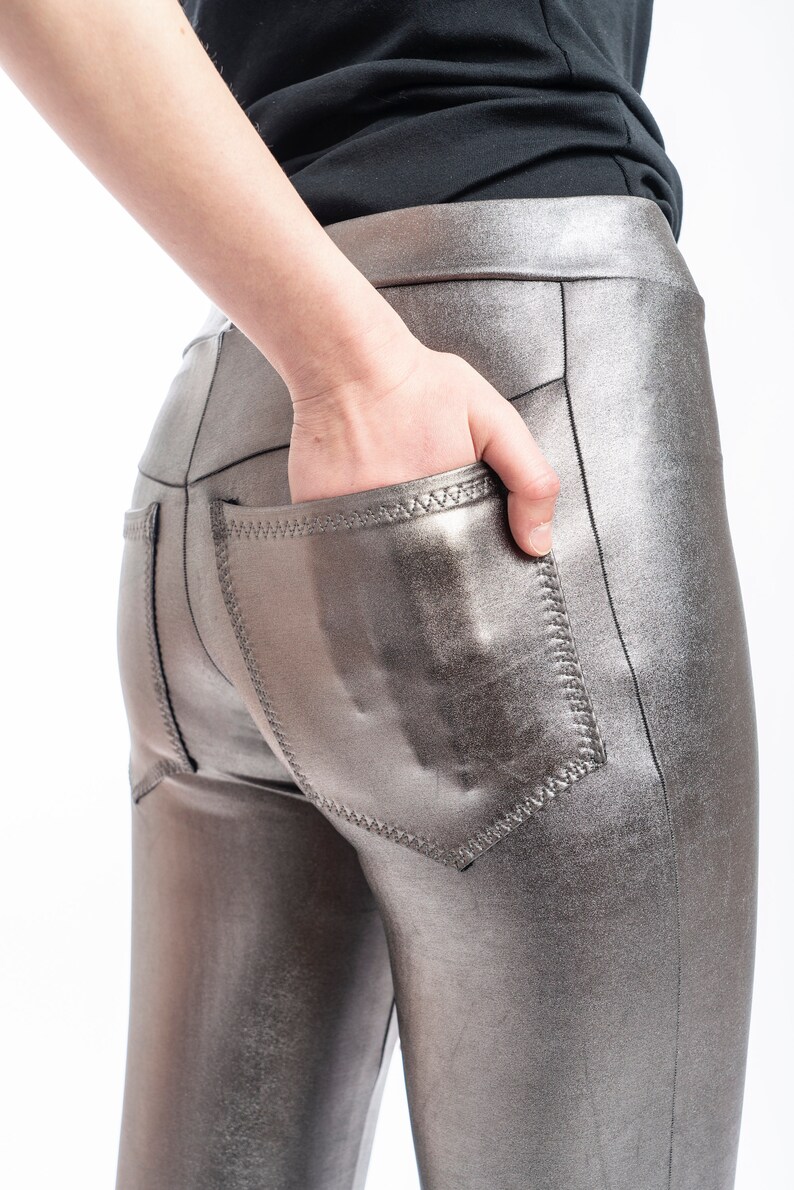 May include: Metallic silver leggings with a high waist and back pockets. The leggings have a shiny, reflective finish and are made of a stretchy material. The back pockets are outlined with a contrasting stitch.