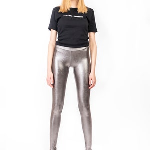 May include: A black t-shirt with the text "SERA DUST" in white, paired with metallic silver leggings and black high heels. The leggings have a shiny, textured appearance.