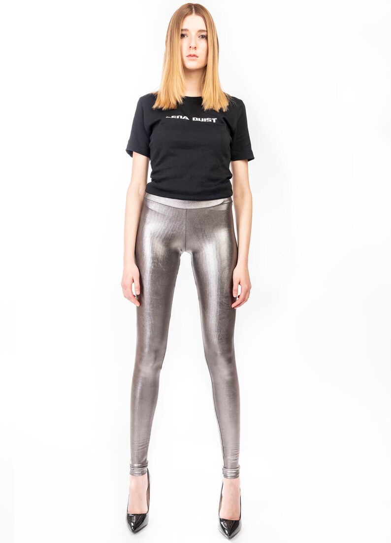 May include: A black t-shirt with the text "SERA DUST" in white, paired with metallic silver leggings and black high heels. The leggings have a shiny, textured appearance.