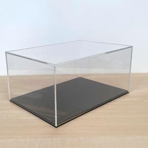 May include: A clear acrylic display case with a rectangular shape. The case has a black base, designed to showcase collectibles. The transparent design allows full visibility of the contents. Dimensions are in centimetres.