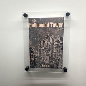 May include: A framed poster of the Hollywood Tower Hotel, with the text "The Hollywood Tower Hotel" and "Drop In!". The poster is in monochrome, showing a stormy night scene with lightning and the hotel.
