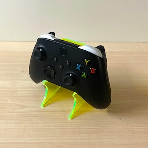 May include: A black games controller with coloured buttons sits on a neon green stand. The controller has white accents and a variety of coloured buttons, including blue, red, green, and yellow. The stand is made of a transparent material.