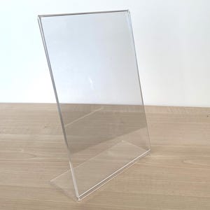 May include: A clear acrylic sign holder with a vertical orientation and a slanted back support. The display stand features a rectangular frame designed to hold paper or cardstock inserts securely in place for tabletop viewing and presentation.