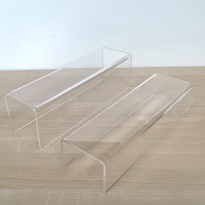 May include: Two clear acrylic risers, each featuring a rectangular top surface and two side panels bent at right angles to form an open-ended bridge structure. These transparent display stands are designed with smooth edges and a minimalist, open design.