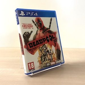 May include: A PS4 video game case for Deadpool, featuring the character in a red and black suit with swords. The case has the text "THIS IS WHAT AWESOME LOOKS LIKE" and "DEADPOOL". The game is rated 18.