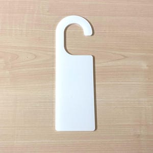 May include: A white door hanger with a curved top and a rectangular base. The hanger is made of a solid, opaque material and is set against a light brown wooden surface.