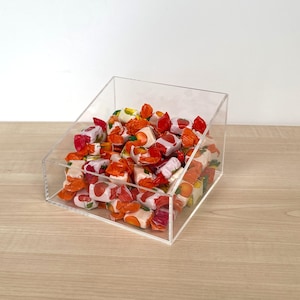 May include: A clear acrylic bin with an angled front opening filled with assorted sweets wrapped in colourful red, orange, and white wrappers. The container is square-shaped and transparent, allowing a full view of the wrapped sweets inside.