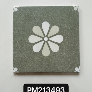 Op de afbeelding: Vierkante decoratieve tegel met een bloemenontwerp. De tegel heeft een gespikkelde, grijsgroene achtergrond met een witte en lichtbeige bloem in het midden. Witte bloemblaadjes accenten bevinden zich in elke hoek. De tekst "PM213493" staat in een zwarte rechthoek.