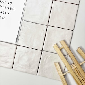 以下が含まれることがあります： オフホワイトの正方形のタイルが格子状に配置されています。木製のイーゼルが右側に配置されています。左側には、黒い文字が書かれた白いカードが部分的に見えています。テキストは「WHAT IS WISHES REALLY YOU.」と書かれています。