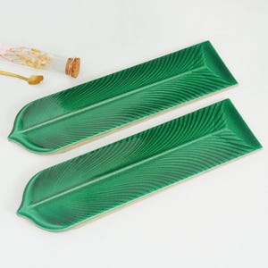 Puede incluir: Dos fuentes verdes de cerámica con forma de hoja. Cada fuente tiene un acabado brillante y veteado detallado. Las fuentes son rectangulares con un extremo redondeado en forma de hoja. El fondo es blanco.